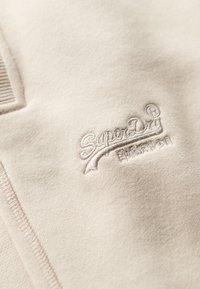 Tissu de sweat-shirt beige avec une texture douce, présentant un logo "SuperDry" brodé dans une couleur de fil complémentaire et des détails en côte.
