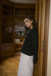 Svart jacka med rund halsringning, knappdetaljer och långa ärmar i kombination med en ljusfärgad kjol med paljetter. Bakgrund i träpanel.