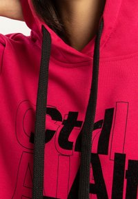 Zbliżenie na osobę w jasnoróżowym dresie z czarnymi sznurkami i częściowo widocznym pogrubionym czarnym tekstem z przodu.