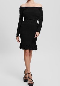 Robe noire à épaules nues avec des rayures horizontales texturées, corsage ajusté et ourlet en forme de vagues. Assortie à des chaussures à plateformes noires à brides.