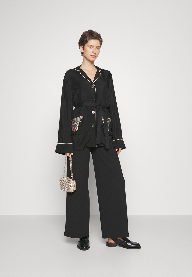 Zwarte satijnen stijljas met knoopsluiting, witte piping en geborduurde details op de zakken, gecombineerd met zwarte wijde broek en een kralen tas.