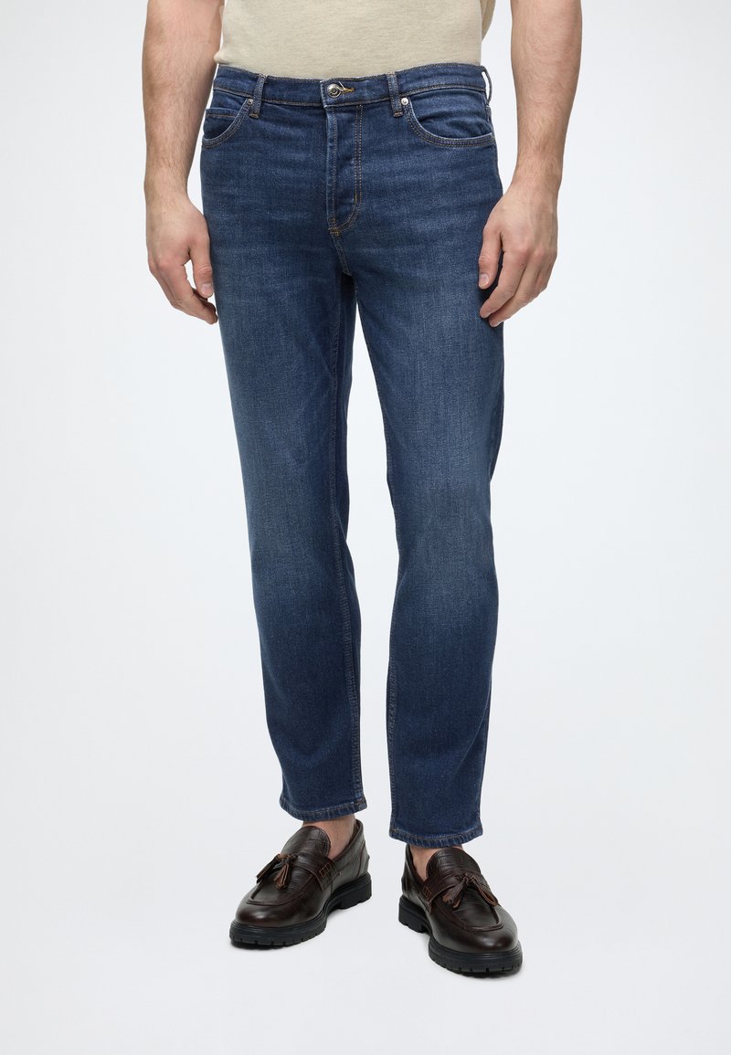 Homme portant un jean slim bleu foncé avec fermeture par bouton et fermeture éclair, associé à des mocassins à pampilles en cuir marron foncé.