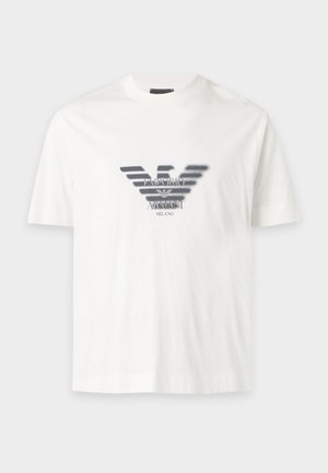 Mann stehend, trägt ein weißes T-Shirt mit schwarzem Logo, dunkle Jeans und schwarze Schuhe vor einem schlichten weißen Hintergrund.