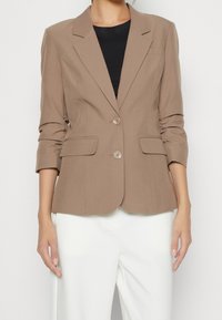 Brun skräddarsydd kavaj med spetsiga slag, två knappar och lockfickor. Mjuk tyg med lätt lyster, figursydda ärmar och en slät textur.