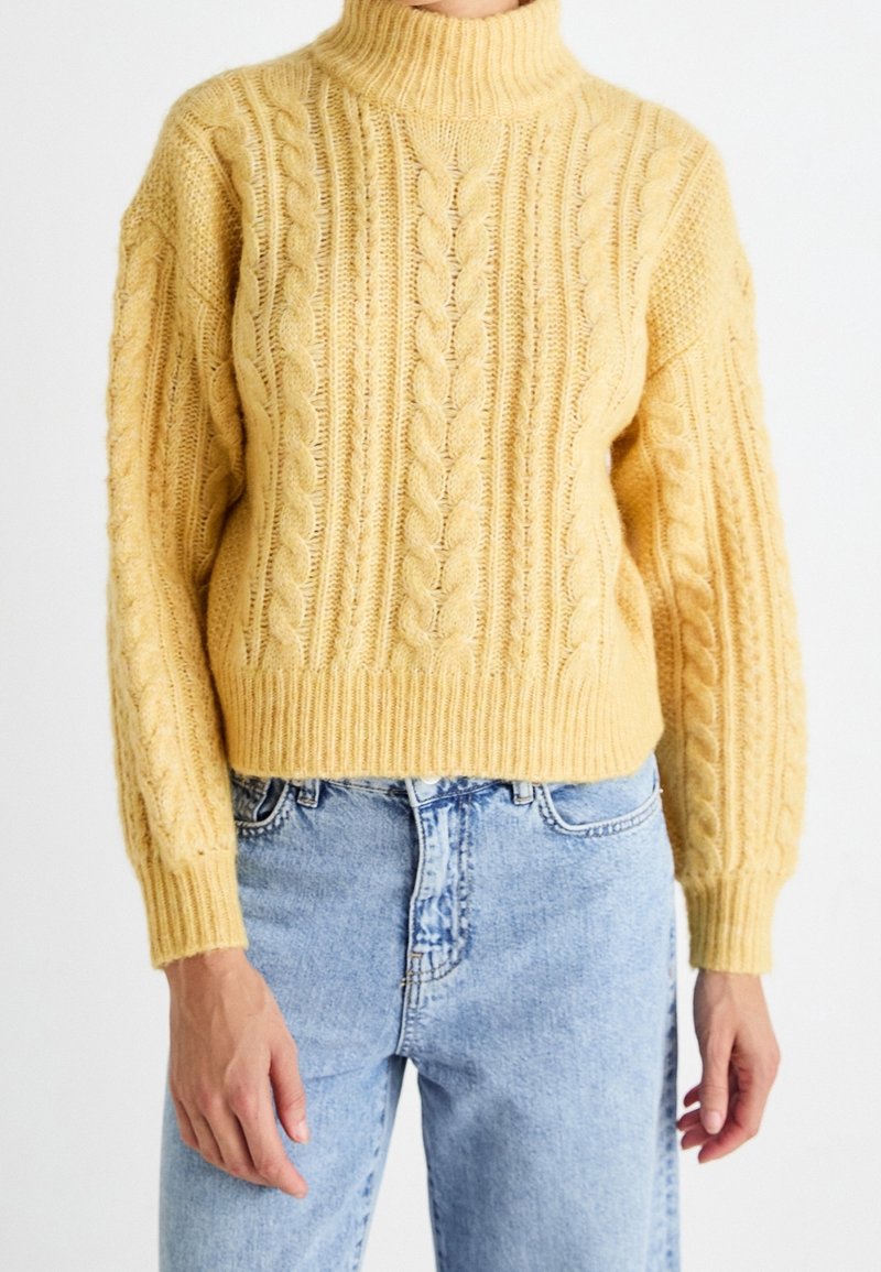 Pull jaune en maille torsadée avec un col montant côtelé et des épaules tombantes. Poignets et ourlet côtelés. Associé à un jean en denim bleu clair.