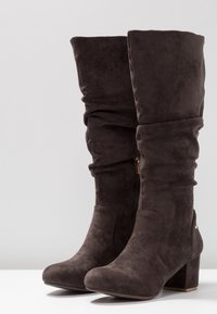 Botas de ante marrón oscuro de altura hasta la rodilla con un diseño arrugado, punta redonda y tacón ancho; cuentan con una cremallera dorada en el lado interno.