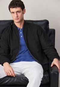 Jeune homme assis sur une chaise en cuir noir, portant une veste zippée noire sur une chemise bleue et un pantalon blanc, regardant légèrement vers la caméra.
