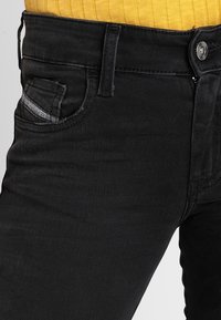 Vaqueros de mezclilla negra con una textura suave, que cuentan con un bolsillo frontal y un cierre de botón metálico. La cinturilla está elevada por encima de una blusa amarilla de canalé.