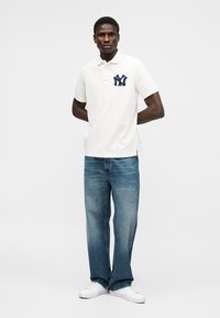 Mężczyzna stoi ze złożonymi za plecami rękami, ubrany w białą koszulkę polo z granatowym logo, niebieskie dżinsy i białe trampki na jednolitym tle.