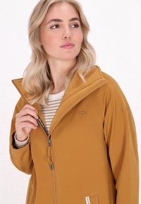 Gelsvai geltona striukė su lygiu paviršiumi, aukštu apykakle, užtrauktuku ir siuvinėtu skėčio logotipu. Po ja matyti dryžuotas marškinėliai.