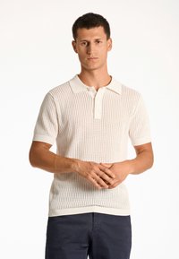 Kortärmad stickad polo skjorta i ljusbeige med en strukturerad öppen mönster och ribbad krage och fåll. Bärs med mörkblå byxor.