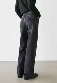 Tmavě modré džínové kalhoty s volným střihem, s kapsami na zádech a hladkou texturou. Modelka má na sobě černý top a rukama si drží kapsy.