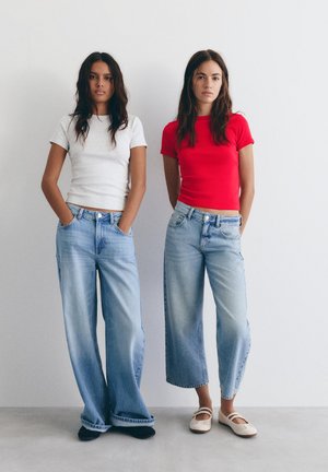 Două femei stând lângă un perete alb, purtând blugi deschisi la culoare și tricouri cu mânecă scurtă; una albă, cealaltă roșie.