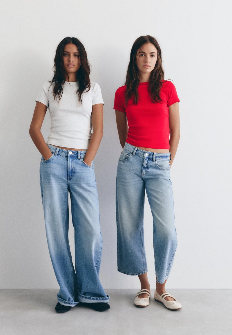 Dwie kobiety stojące przy białej ścianie, ubrane w jasnoniebieskie dżinsy i koszulki z krótkim rękawem; jedna w białej, druga w czerwonej.