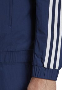 Marineblaue Sportjacke mit weißen Streifen an den Ärmeln, ausgestattet mit elastischen Bündchen und einem strukturierten Stoff. Angenehmer Schnitt mit Kordelzug.