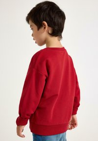 Sweatshirt rouge en coton avec des manches longues légèrement surdimensionnées et un col ras du cou, associé à un jean bleu clair. Vue de dos.