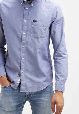Homme portant une chemise bleu clair à manches longues avec une poche poitrine et un jean bleu clair, une main dans la poche et l’autre le long du corps.
