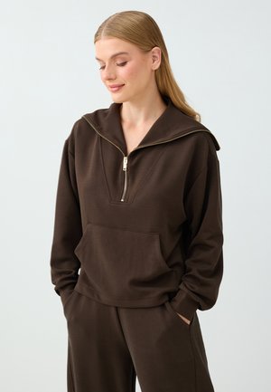 Un mannequin porte un ensemble d’intérieur côtelé marron foncé, composé d’un pull à fermeture éclair et à col large et d’un pantalon assorti, sur un fond clair.