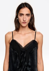 Czarna koszulka na ramiączkach z koronkową lamówką i satynową fakturą. Posiada cienkie, regulowane ramiączka oraz ozdobną kokardę z przodu. Prosty, elegancki design.