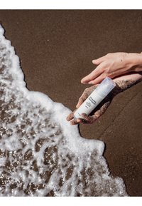 Des mains couvertes de sable tiennent un flacon blanc de crème solaire près des vagues mousseuses de l'océan déferlant sur une plage de sable.
