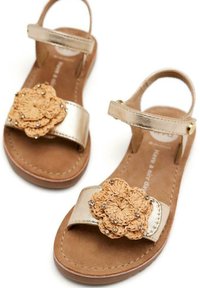 Sandalias metálicas doradas con un acento de ganchillo floral en tono marrón y adornos de strass sobre una plantilla de texturizada marrón. Correa ajustable en el tobillo.