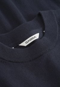 Pulover de culoare albastru marin, cu guler rotund cu model în relief și tricot moale, texturat. Eticheta este vizibilă, având inscripția "GABBA" în negru pe fundal alb.
