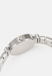Montre en acier inoxydable argenté avec un boîtier rond, texture lisse, détail de couronne et bracelet en métal maillé. Design minimaliste, sans logo visible.