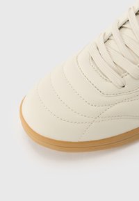Sneaker en cuir crème avec bout rond, tige texturée et semelle en caoutchouc beige. Doté de lignes cousues et de lacets plats.