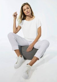 Top corto bianco a maniche corte e pantaloni chiari, abbinati a sneakers bianche. Il modello posa seduto su un pouf grigio testurizzato. Sfondo luminoso.