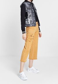 Denimväst med mönstrade detaljer, svart långärmad topp, korta gula byxor med broderi, och vita sneakers med blommönster.