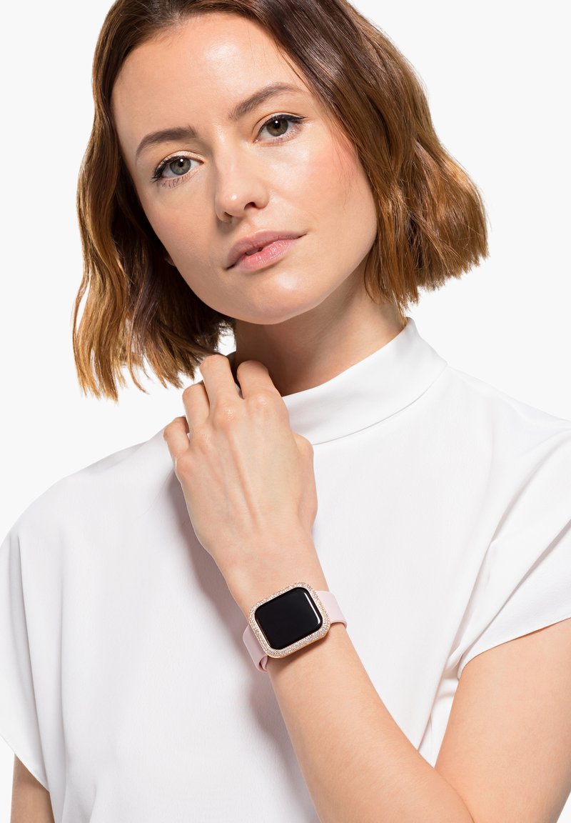 Jonge vrouw met kort bruin haar draagt een witte top en een roze smartwatch met een glinsterend frame aan haar linkerpols.