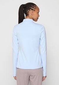 Maglia a maniche lunghe azzurro chiaro con texture a coste, colletto alto e orlo dritto, abbinata a pantaloni chiari. Nessun logo o motivo visibile.