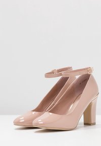 Ett par glänsande nakena rosa högklackade skor med ankelremmar och guldspänne, utställda på en vit yta mot en enfärgad bakgrund.