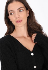 Zwarte gebreide cardigan met een V-hals, textuurpatroon en zilveren knopen. Zacht materiaal met lange mouwen.