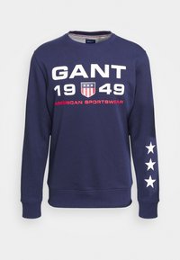 Navyblauw sweatshirt met wit "GANT" logo en rode accenten, met een schild-embleem, het jaar "1949" en witte sterren op de mouwen.
