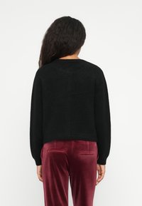 Femeie cu păr lung și închis la culoare, purtând un pulover negru și pantaloni din catifea burgundă, stând cu spatele la un fundal alb simplu.