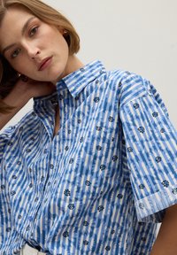 Camicia a maniche corte blu e bianca a righe con motivi floreali. Presenta un colletto con bottoni e una tasca sul petto. Texture del tessuto morbida.