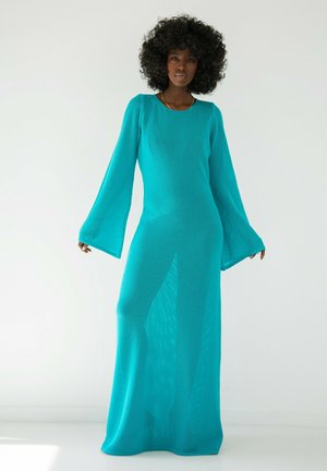 Długa, turkusowa dzianinowa sukienka z dzwonowymi rękawami i luźnym krojem. Przezroczysta faktura, sięgający do podłogi fason oraz okrągły dekolt.