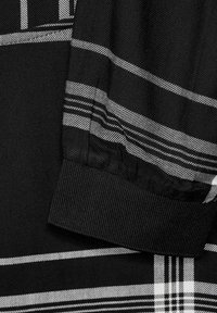 Tessuto nero con motivo a quadri grigi. Presenta polsini elasticizzati e una texture liscia. Le linee di accento sono sottili e strutturate.