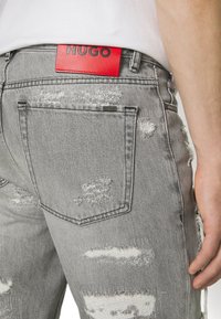 Harmaat kuluneet farkut takataskulla ja punaisella "HUGO" -tunnuksella, yhdistettynä valkoiseen teeppariin, näyttäen osan henkilön kädestä ja käsivarresta.