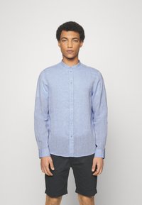 Camisa de botones de color azul claro con cuello de banda, con mangas largas y una tela texturizada. Combinada con pantalones cortos oscuros.