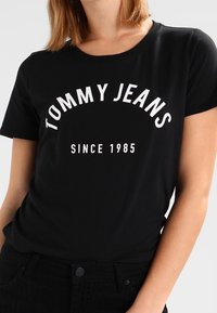 Svart bomull T-shirt med texten "TOMMY JEANS" i vitt i en böjd stil, med "SINCE 1985" nedanför i rak linje. Kort ärm och rundad nacke.