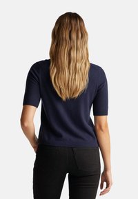 Žena s dlouhými vlnitými blond vlasy, oblečená v námořnickém svetru s krátkým rukávem a černými kalhotami, stojí zády k bílé pozadí.