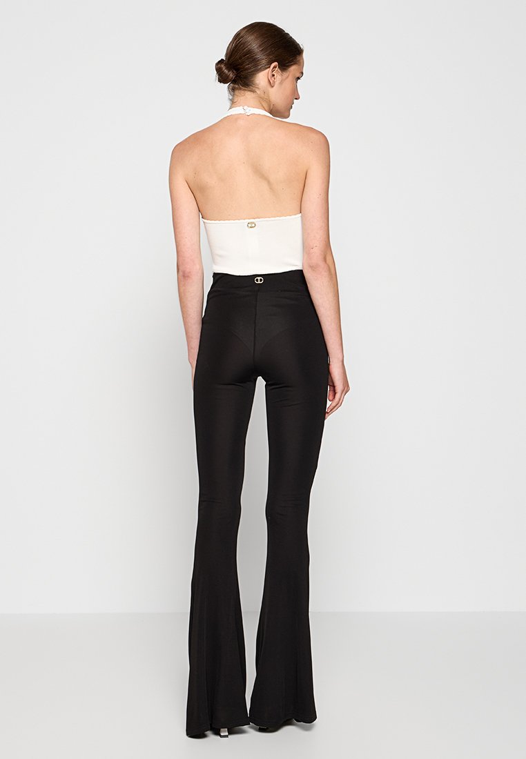Pantalones negros acampanados de cintura alta, combinados con una blusa sin mangas blanca que presenta un escote halter y un detalle de botón dorado en la parte de atrás.
