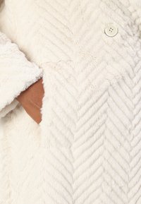 Weißer Stoff mit Fischgrätmuster, versehen mit einem Knopfdetail. Die Textur wirkt weich und plüschig und vermittelt ein warmes Aussehen.