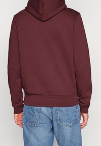 Sweat à capuche bordeaux avec une grande capuche, des poignets et un ourlet côtelés. Le tissu semble doux et texturé. Porté avec un pantalon en denim bleu ample.