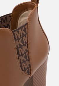 Bottine en cuir marron à talon haut avec panneau latéral élastique présentant un motif répété du logo "MK" en marron foncé et beige.