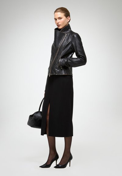 Nainen mustassa nahkatakissa, mustassa midi-hameessa, jossa halkio, läpikuultavissa sukissa, mustissa korkeakoroisissa kengissä, mustaa käsilaukkua kantaen, seisomassa yksinkertaisen taustan edessä.