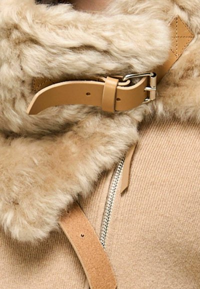 Cappotto in shearling beige con colletto in morbido pelliccia, cinturino in pelle marrone chiaro con fibbia e zip in argento visibile su tessuto testurizzato.