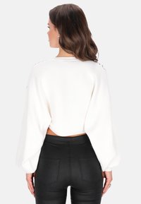 Witte geribbelde crop trui met ballonmouwen, blootgesteld achterpand en decoratieve knoopaccenten op de schouders, gecombineerd met zwarte strakke broek.